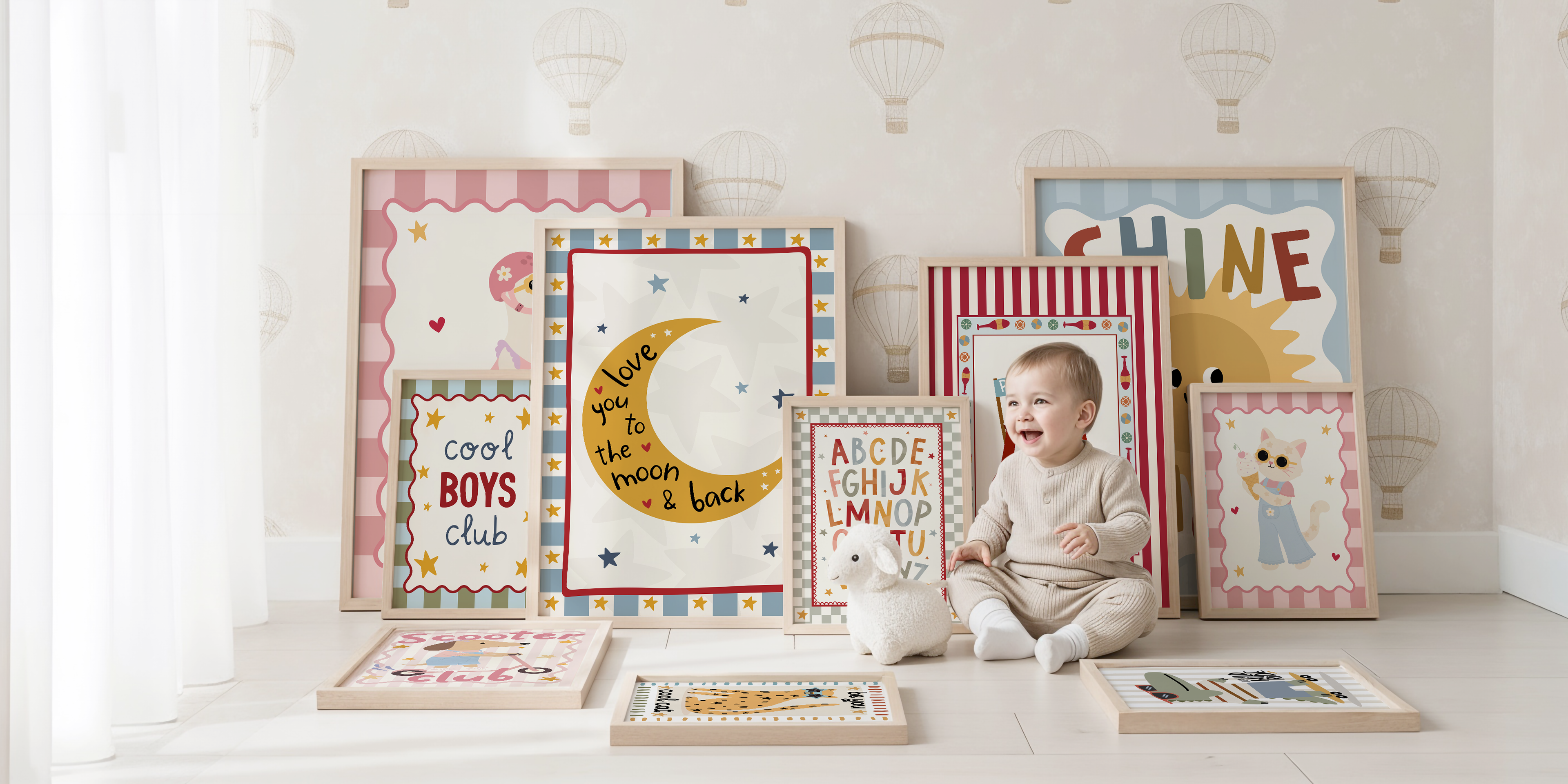 A baby in a cream outfit stands among a group of framed nursery art prints and decorative boards in a bright, white room. The prints feature pastel patterns, a crescent moon, alphabet letters, and playful text including 'cool BOYS club' and 'shine'.
